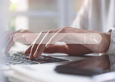 Hands typing on a laptop keyboard with a transparent search bar overlay and magnifying glass icon, symbolizing online search and digital work.
